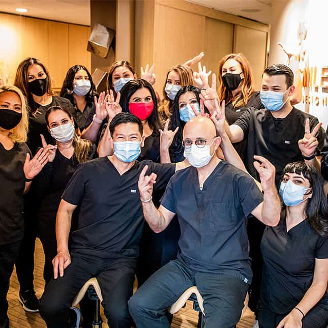 Dental team members wearing masks and dark scrubs waving at camera in office setting.