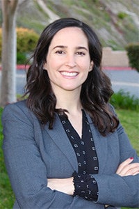 Professional woman smiling at camera, arms crossed, outdoor setting with blurred background.