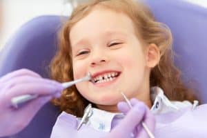 Young girl with reddish-brown hair smiling brightly in dental chair wearing purple bib.