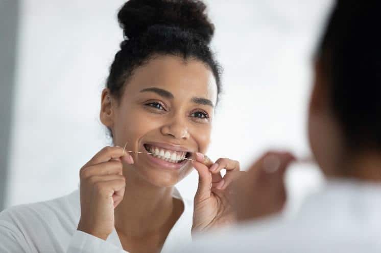 Woman smiling while flossing teeth in bathroom mirror during morning dental routine.