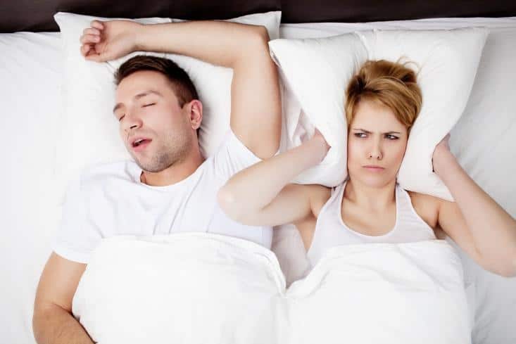 Couple in bed with relationship tension, man sleeping while woman appears frustrated or concerned.