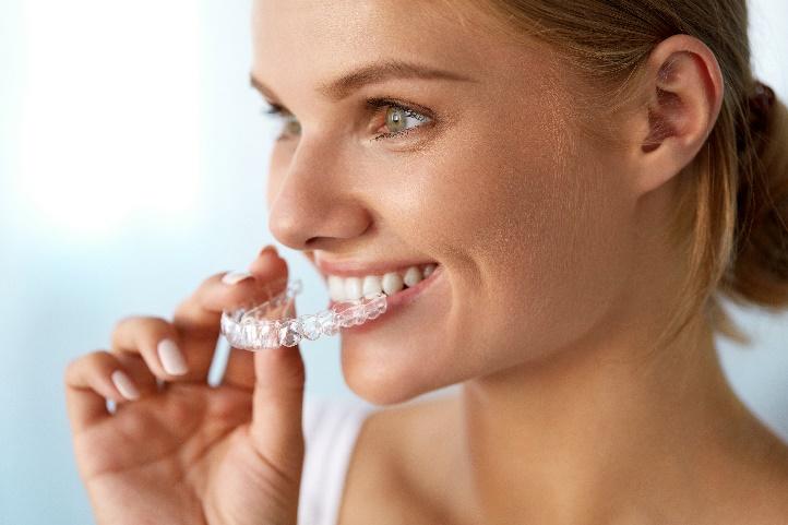 Woman holding clear aligner tray against her teeth, smiling against white background.