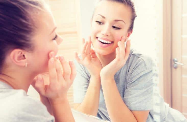 Woman applying skincare product to face while smiling at bathroom mirror reflection