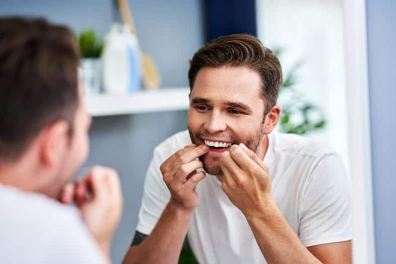 Man examining teeth in bathroom mirror while brushing and flossing daily.