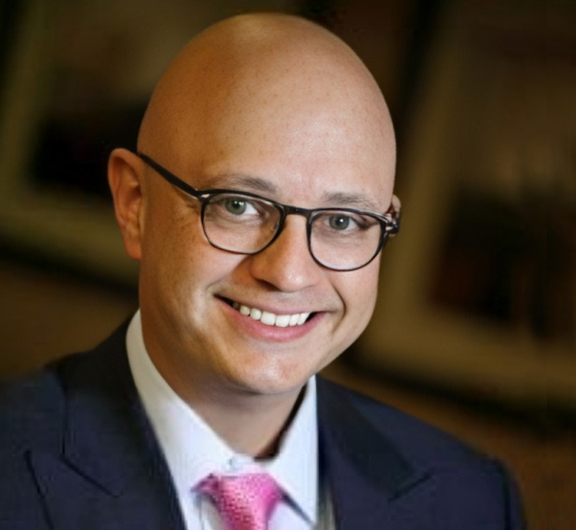 Professional headshot of bald man wearing glasses and pink tie smiling at camera