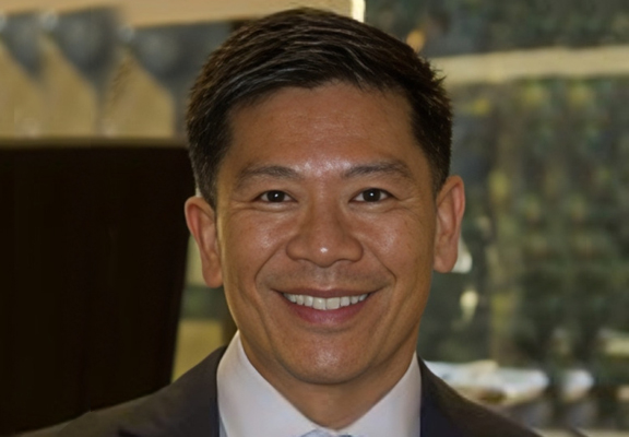 Professional headshot of smiling man in business suit against blurred office background.