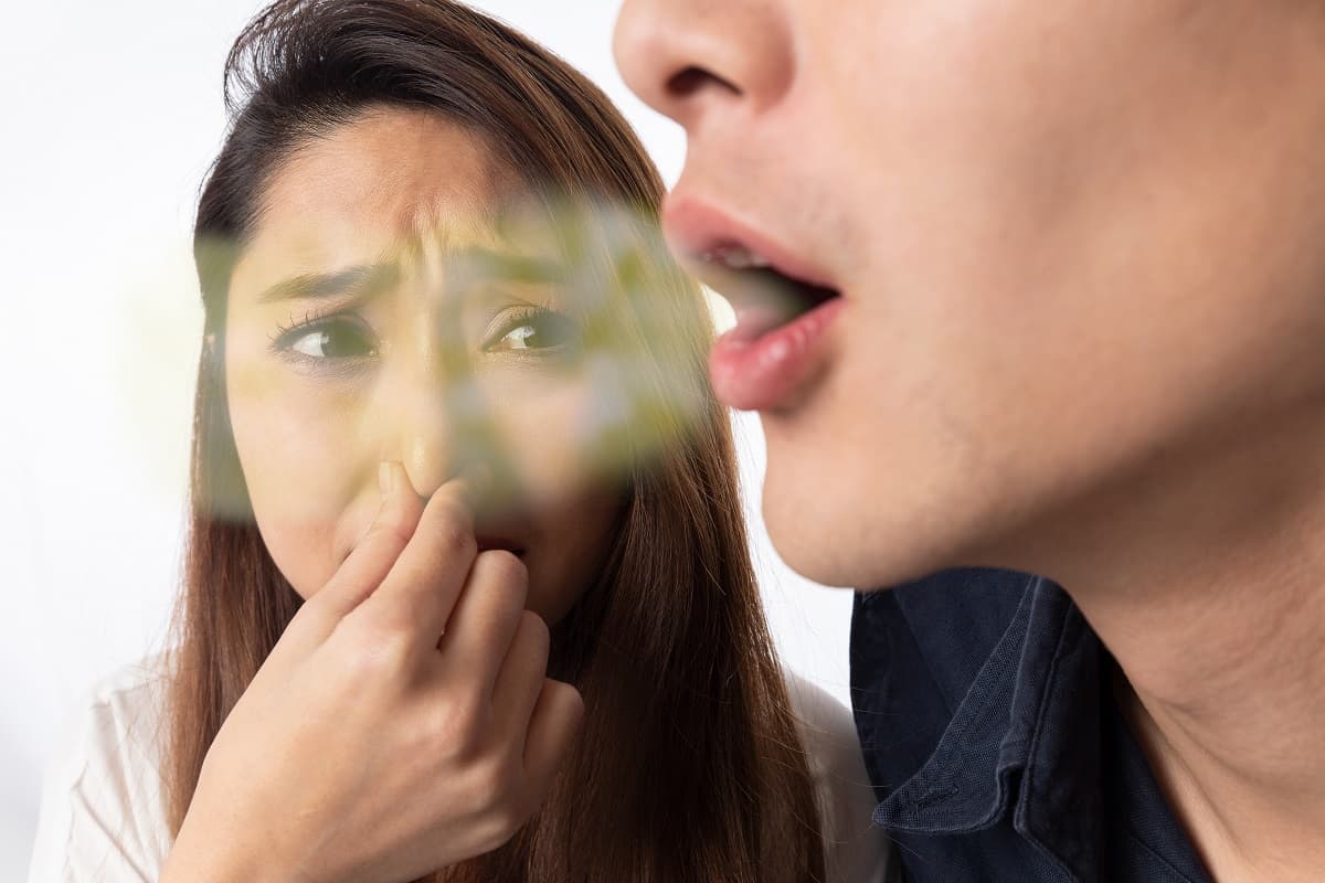 Man exhaling bad breath toward concerned woman with hand near mouth indoors.