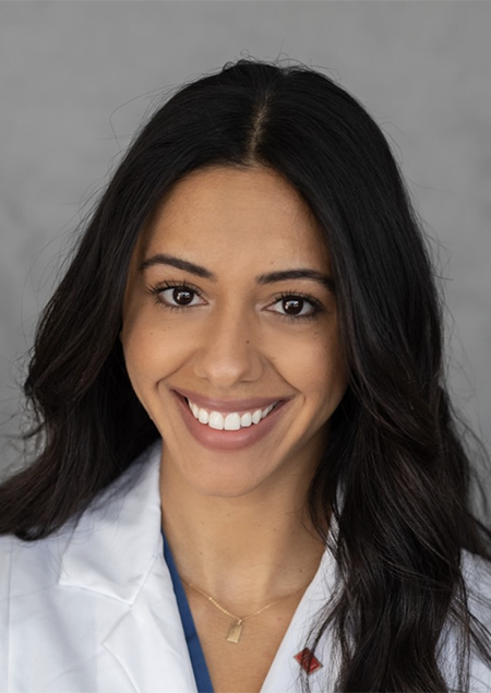 Professional headshot of female dentist in white coat smiling at camera against gray background.