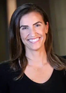 Woman smiling at camera, professional headshot, black top, indoor setting with neutral background.